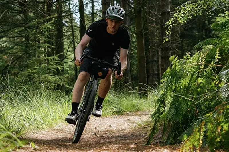 Ridiang a gravel bike in the woods on a sunny day wearing an MTB style helmet